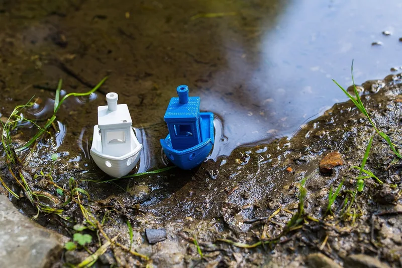 Dois barquinhos Benchy impressos em 3D, um branco e um azul, boiando na beira de uma poça